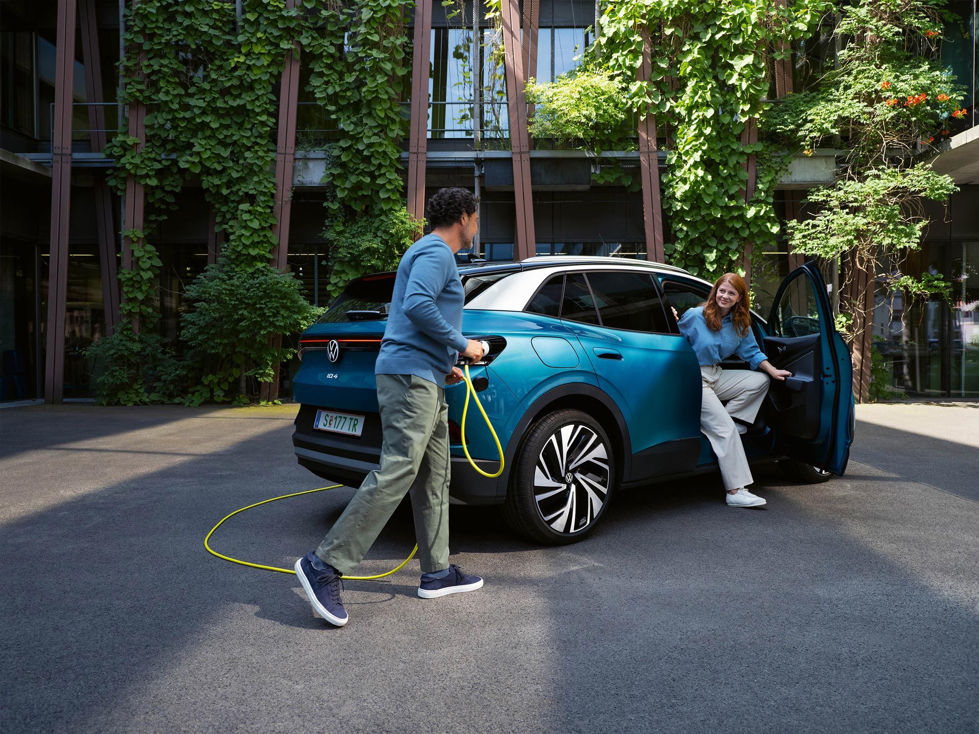 Ein Mann mit einem gelben Ladekabel für Elektroautos geht auf das Heck eines parkenden blauen VW ID.4 zu. Aus der Beifahrertür steigt eine junge Frau aus.