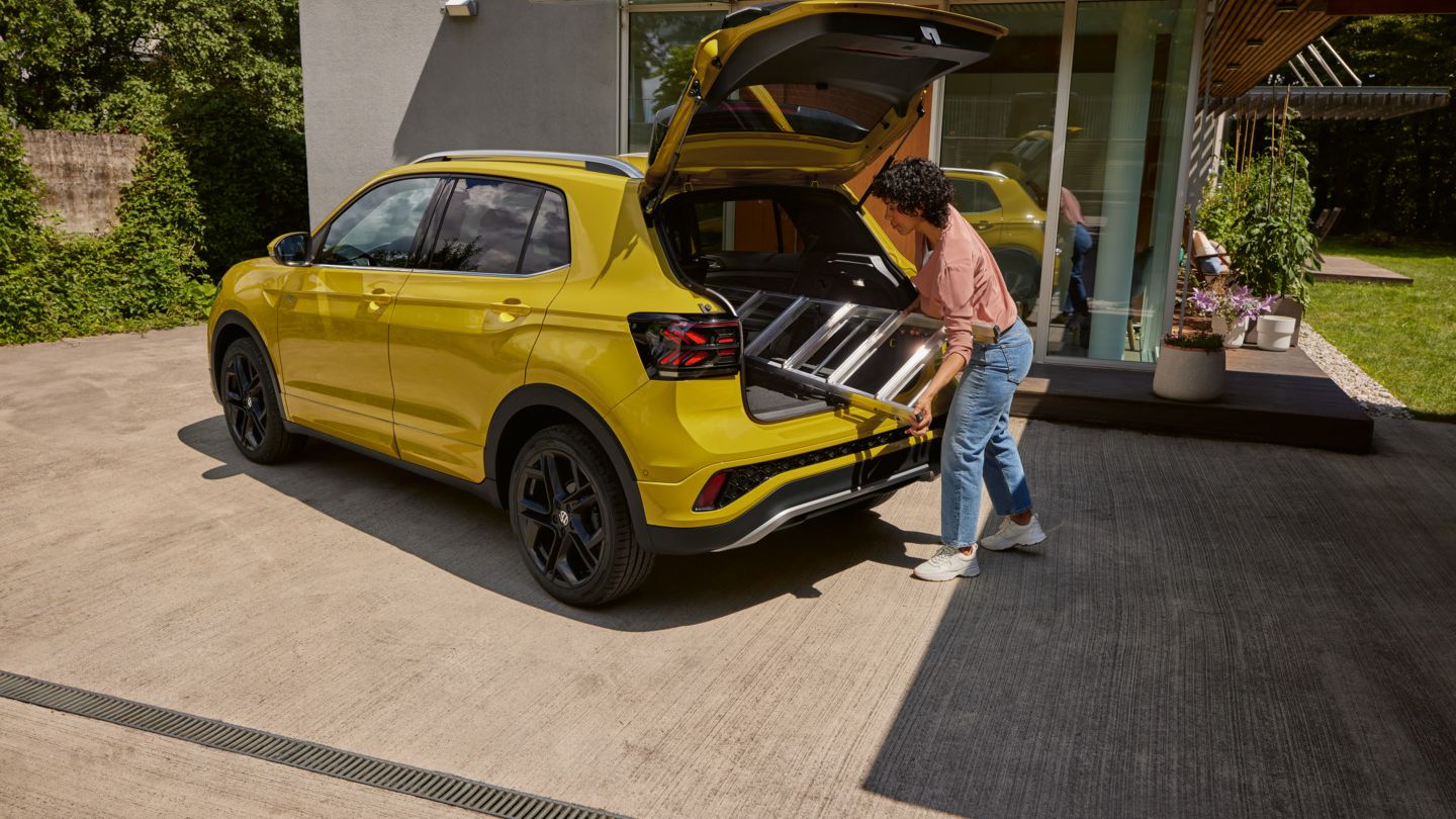 Eine junge Frau schiebt eine Leiter in den Kofferraum eines gelben VW T-Cross.
