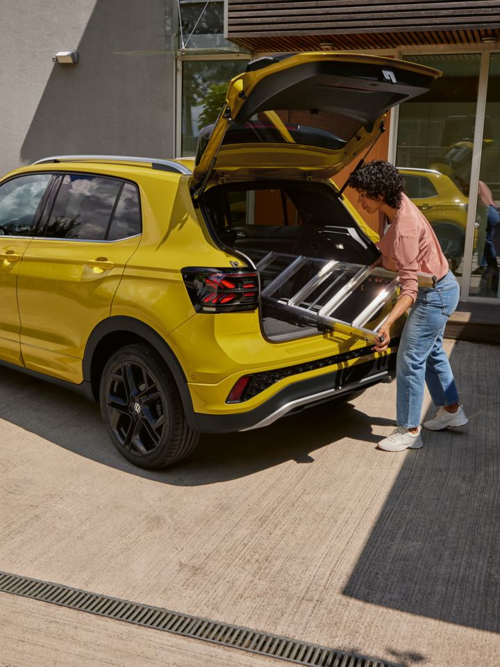 Eine junge Frau schiebt eine Leiter in den Kofferraum eines gelben VW T-Cross.