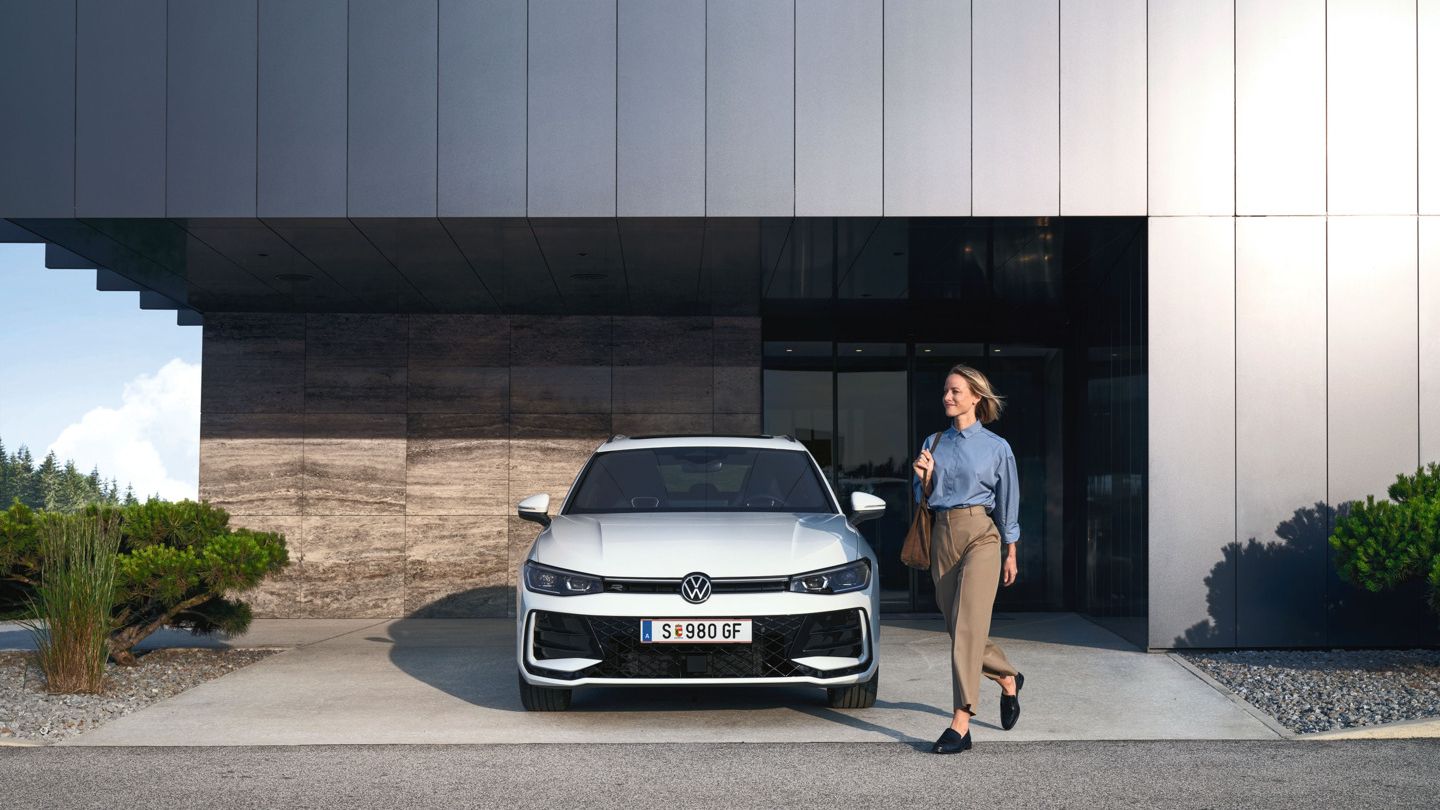 Frontansicht eines weißen VW Passat R-Line, eine Frau geht an ihm vorbei.