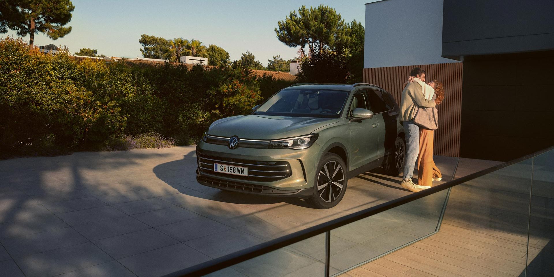 Eine Frau und ein Mann umarmen sich, neben ihnen steht ein VW Tiguan Elegance in der Farbe Cipressino-Grün Metallic