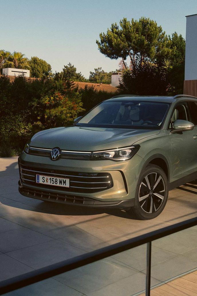 Eine Frau und ein Mann umarmen sich, neben ihnen steht ein VW Tiguan Elegance in der Farbe Cipressino-Grün Metallic