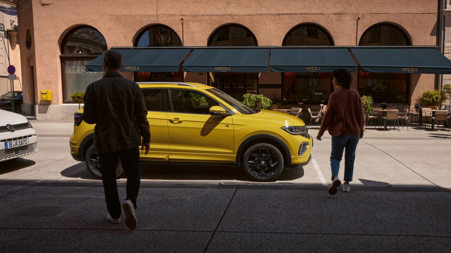 Mann und Frau nähern sich einem gelben VW T-Cross am Straßenrand.