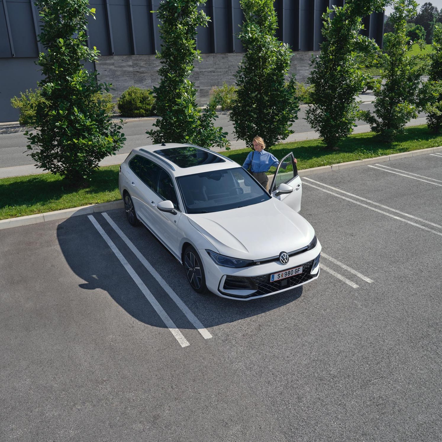 Topshot eines weißen VW Passat R-Line auf einem Parkplatz, eine Frau steigt auf der Fahrerseite ein.