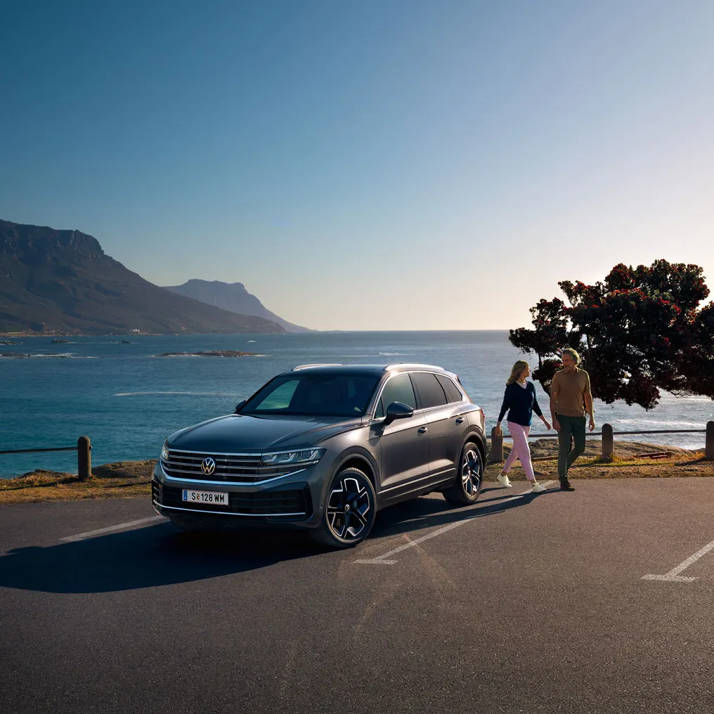 Der neue Volkswagen Touareg auf einem Parkplatz an der Küste, an ihm gehen eine Frau und ein Mann vorbei