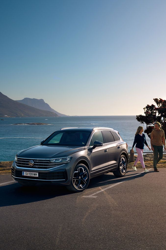 Der neue Volkswagen Touareg auf einem Parkplatz an der Küste, an ihm gehen eine Frau und ein Mann vorbei