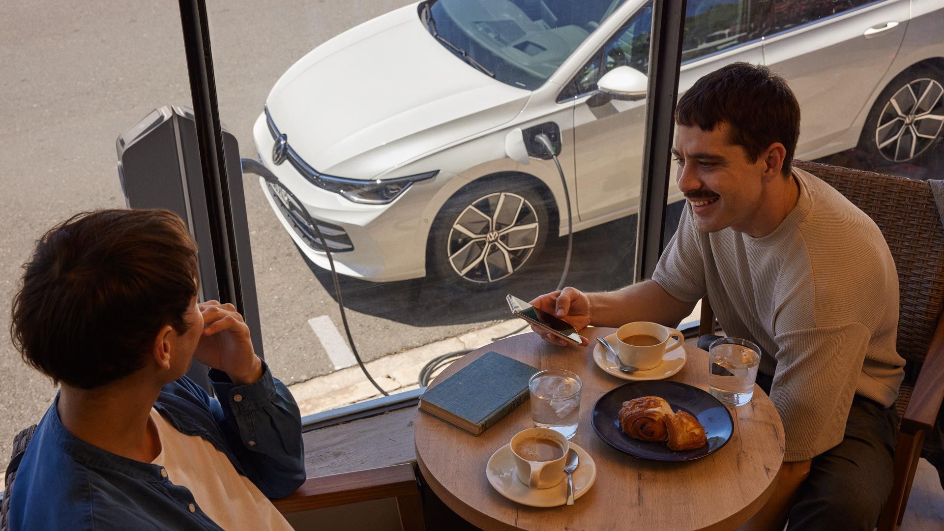 Zwei Männer sitzen in Café und unterhalten sich, durch das Fenster sieht man einen VW Golf eHybrid der geladen wird