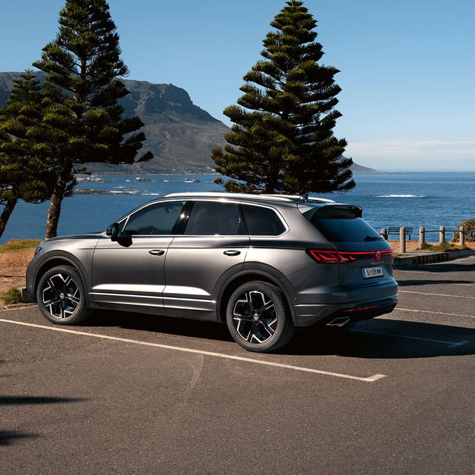 Ein VW Touareg Elegance parkt in der Sonne, Ansicht von der Seite, dahinter sind das Meer und die Küste zu sehen.