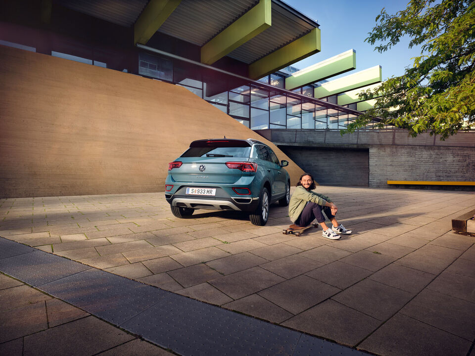 VW T-Roc Style in Blau vor Gebäude geparkt, Heck mit Rückleuchten sichtbar. Man sitzt auf Longboard neben dem Auto.