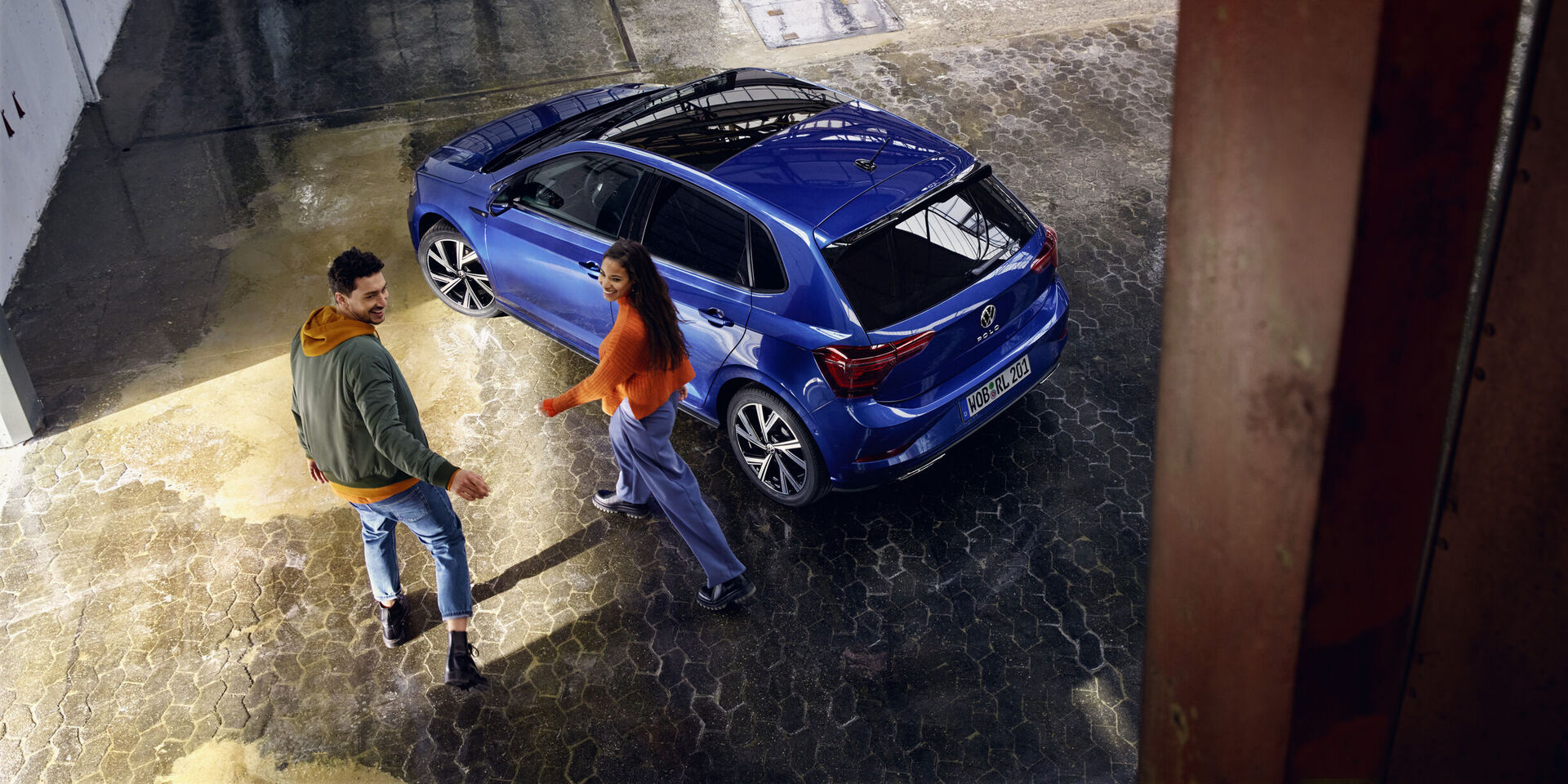 Ein VW Polo in Blau mit optionalem Panoramaschiebedach steht in einer Halle, ein Pärchen geht darauf zu.