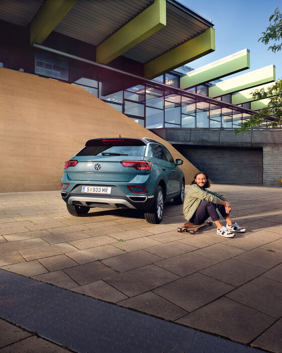VW T-Roc Style in Blau vor Gebäude geparkt, Heck mit Rückleuchten sichtbar. Man sitzt auf Longboard neben dem Auto.