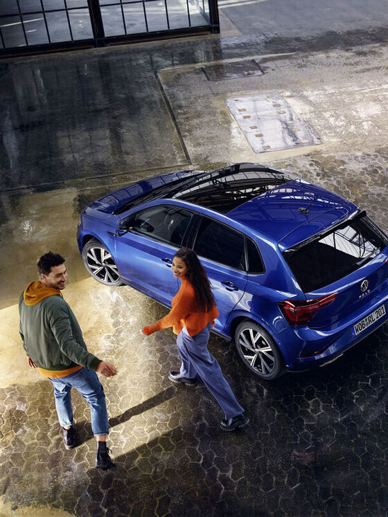 Ein VW Polo in Blau mit optionalem Panoramaschiebedach steht in einer Halle, ein Pärchen geht darauf zu.